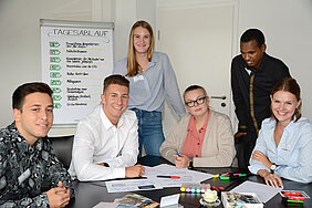 A group photo of the trainees from the year 2019. Six people, including three women and three men, are sitting at a wooden table, smiling at the camera. Papers and writing materials are scattered on the table. Behind them, there is a flipchart displaying the daily schedule.