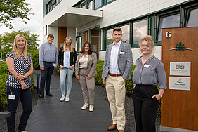 A photo of the trainees from the year 2020. Six people, including four women and two men, are wearing business attire and standing in front of the entrance to the OTG location in Hittfeld, Seevetal. 
