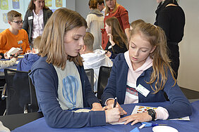 A photo of two girls on the Future Day at LSH, Laurens Spethmann Holding. They are sitting next to each other at a table, wearing name tags. Their gazes are directed downwards as they write something on a piece of paper. Behind the girls, other children can be seen sitting at a table.
