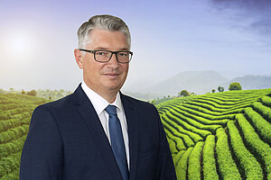 A photo of Ingo Steinbach, a board member of LSH. A man with fair skin, glasses, and short gray hair, wearing a suit with a blue tie. He is standing in front of a hilly landscape of a tea plantation.